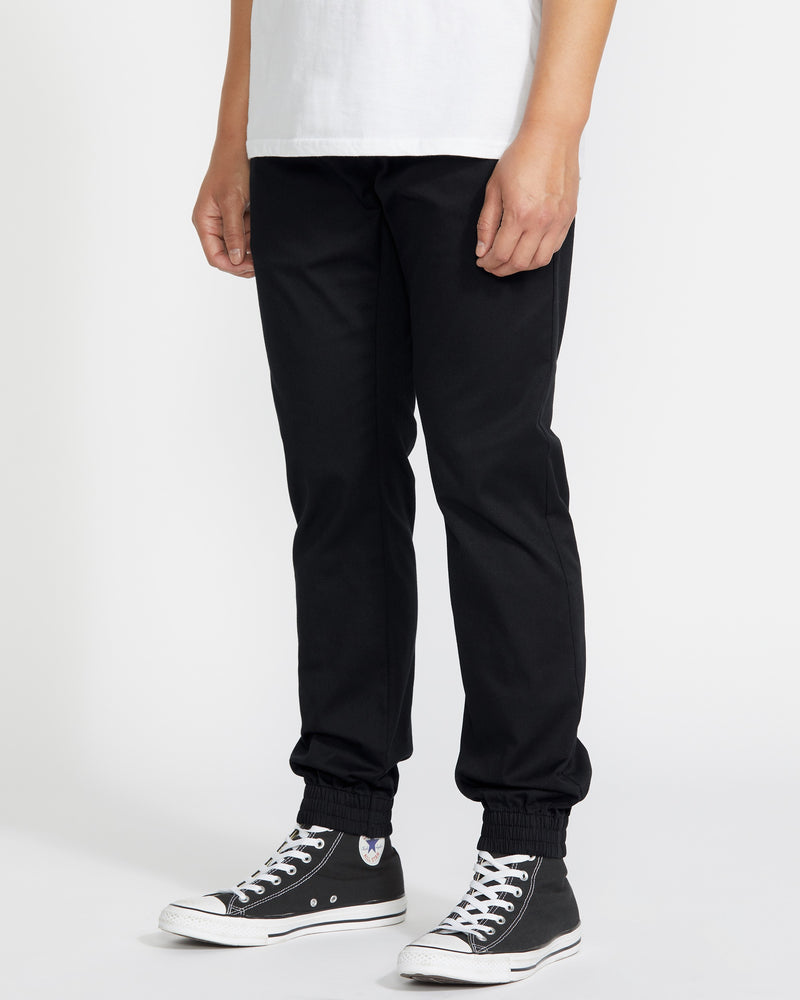A person stands against a plain white background, cropped from shoulder to feet, wearing a white T-shirt and Volcom Men's Frickin Slim Joggers in black, made with REPREVE recycled fibers, plus black high-top Converse sneakers.