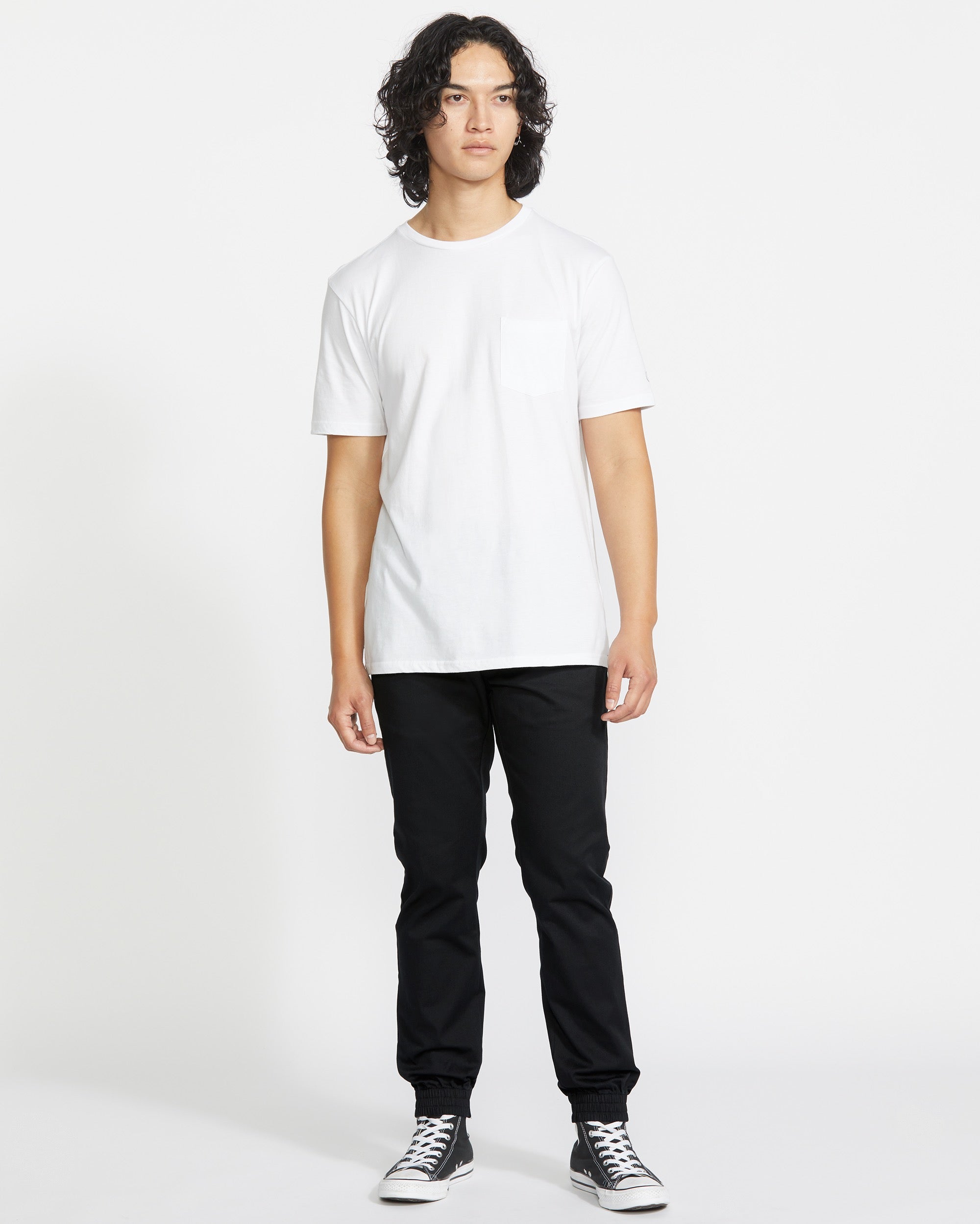 A person with curly dark hair stands facing forward, wearing a plain white t-shirt, Volcom Men's Frickin Slim Joggers in black, and black high-top sneakers against a plain white background.