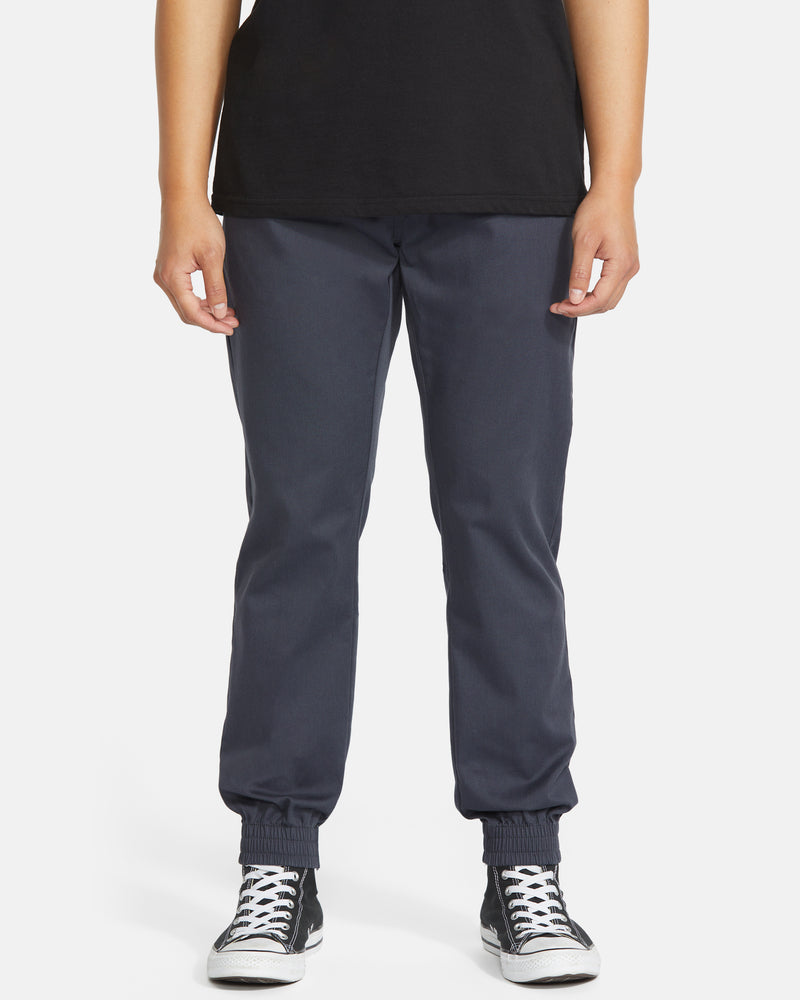 A person wearing a black t-shirt, Volcom Men's Frickin Slim Joggers in dark blue with elastic cuffs, and black-and-white sneakers stands against a plain white background.