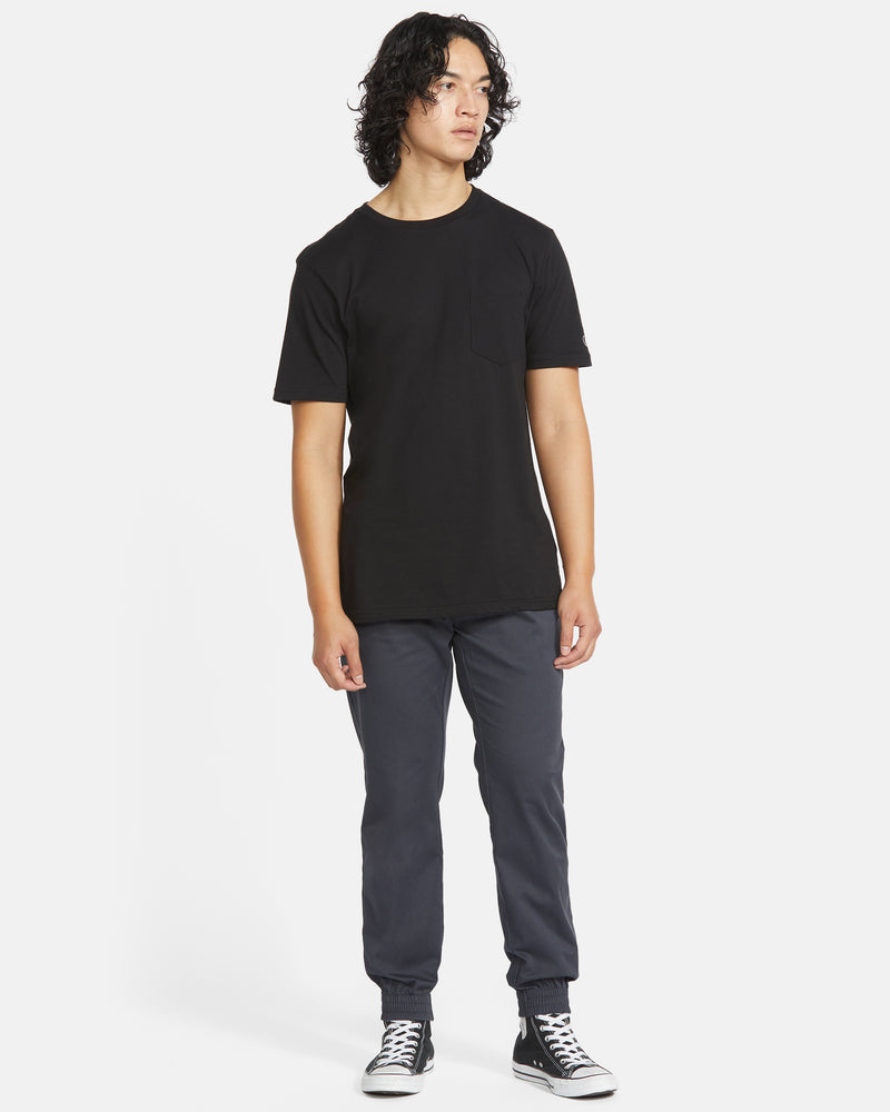 A person with medium-length curly hair stands sideways, wearing a black t-shirt, Volcom Men's Frickin Slim Joggers, and black-and-white sneakers against a plain white background.