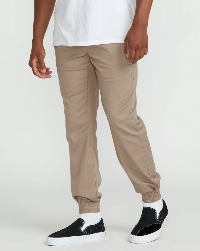 A person wears Volcom Men's Frickin Slim Joggers in tan along with a white T-shirt. Their hands are relaxed at their sides, and they have on black slip-on sneakers with white soles. The background is plain and light-colored.