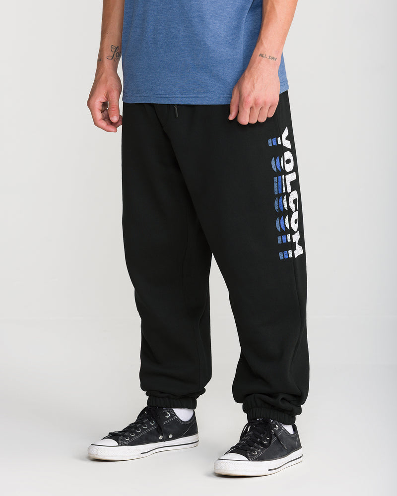 A person wearing Volcom Men’s Quadratic Fleece Pant with an elastic waist, a blue T-shirt, and black sneakers stands against a light background. Only their lower body is visible.