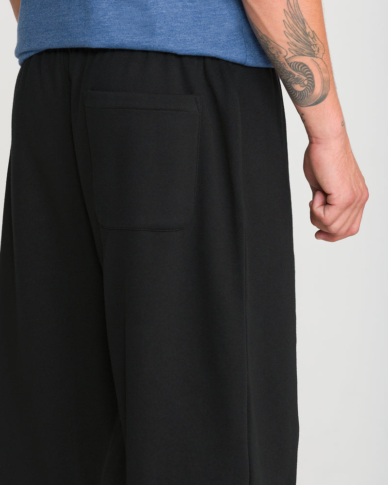 A person is shown from the waist down wearing Volcom Men’s Quadratic Fleece Pant in black with an elastic waist and back pocket. Their right arm, featuring a snake tattoo, is relaxed at their side.