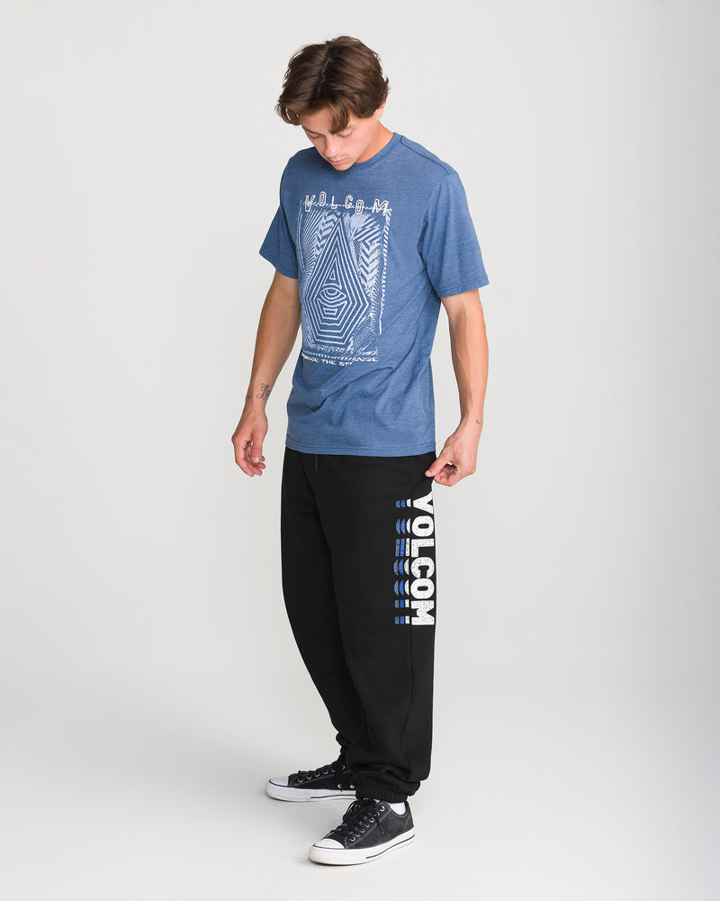 A man wears a blue graphic tee, Men’s Quadratic Fleece Pant by Volcom, and black-and-white sneakers while standing against a plain white background, looking down.
