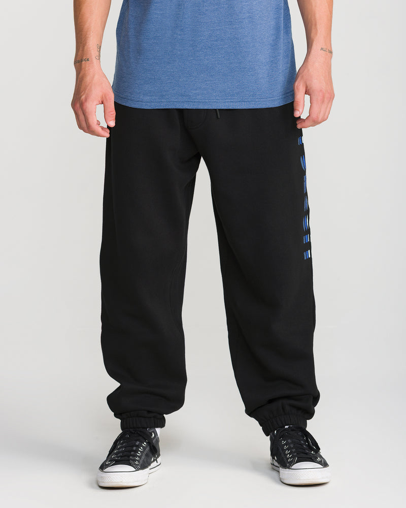 Model wears Volcom Men’s Quadratic Fleece Pant in black with elastic waist and side writing, paired with black sneakers with white soles. Only the lower half is shown, set against a plain background.