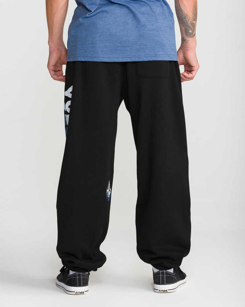 A person wearing a blue T-shirt, Volcom Men’s Quadratic Fleece Pant with graphics and elastic waist, and black sneakers stands with their back to the camera against a plain light background.