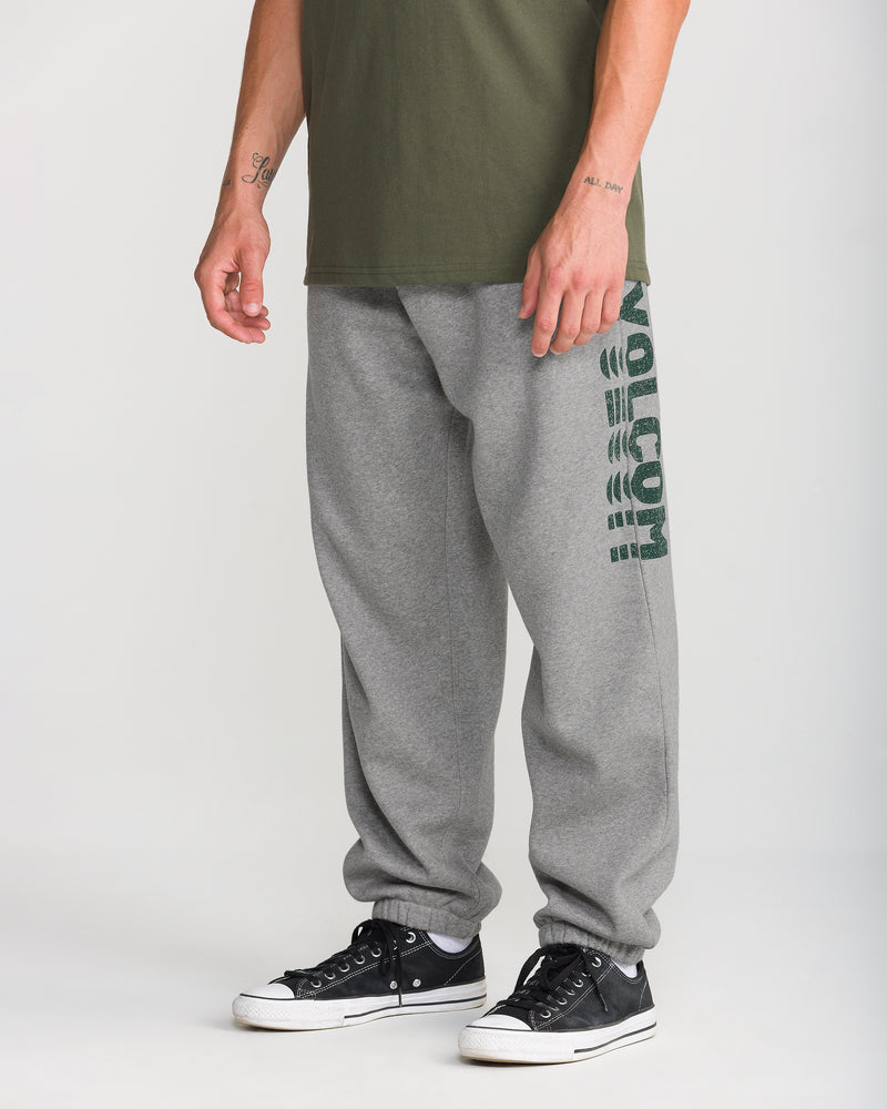A person in Volcom Men’s Quadratic Fleece Pant—classic fit, gray, with VOLCOM on one leg—paired with a green shirt and black sneakers with white soles, stands against a plain background.