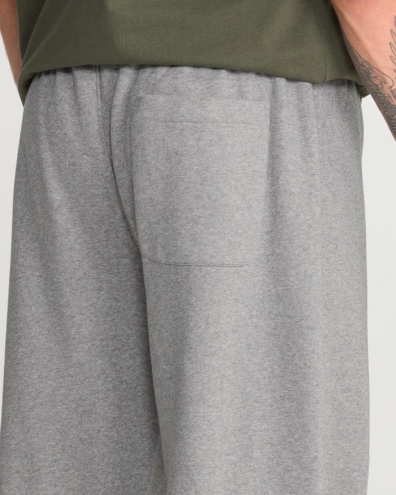 A person wearing Volcom Men’s Quadratic Fleece Pant in gray and an olive green shirt stands with their back to the camera. The classic fit sweatpants feature a back pocket and their arm has a visible tattoo.