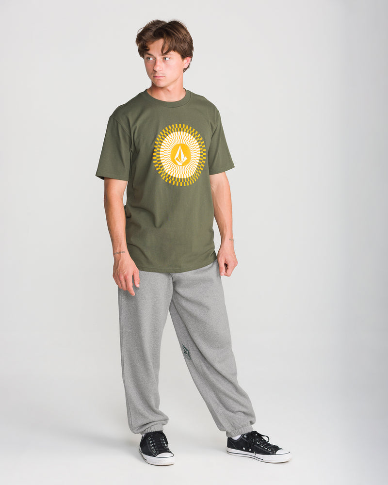 A person wearing a Volcom olive green graphic t-shirt, Men’s Quadratic Fleece Pant in gray, and black sneakers stands against a plain light background, looking to the side with hands relaxed by their sides.