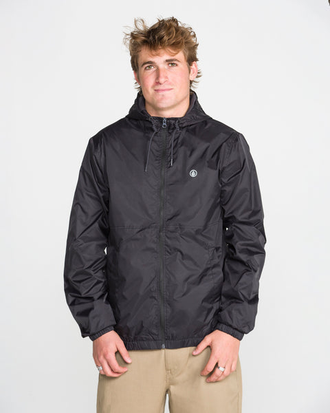 A man with short, tousled hair models the Volcom Men's Ermont Jacket in black, paired with beige pants, standing against a plain white background and looking at the camera with a neutral expression.