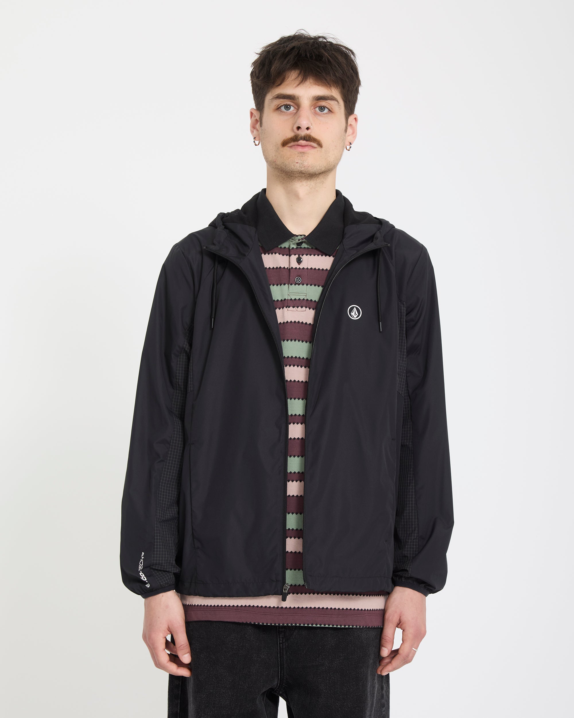 A young man with short hair and a mustache wears the Volcom Men's Wavern Jacket in black over a striped shirt, standing against a plain white background, facing the camera with a neutral expression.