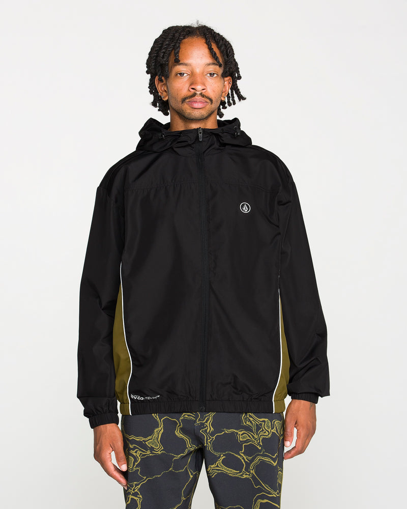A person stands facing forward against a plain white background, wearing the Volcom Men's Wavestone Jacket in black, featuring a mesh-lined hood and white piping, paired with patterned black pants.