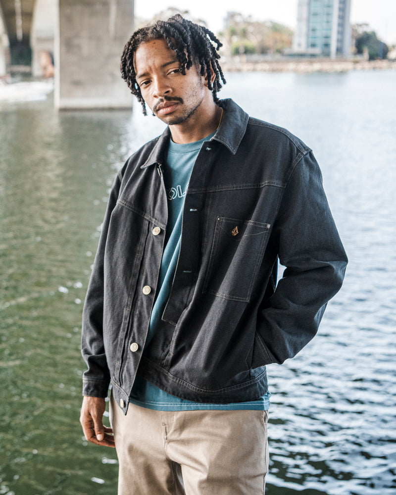 A man with short dreadlocks stands near water under a bridge, with buildings in the background, wearing the Volcom Men’s Slakers Jacket in black, a blue shirt, and beige pants.
