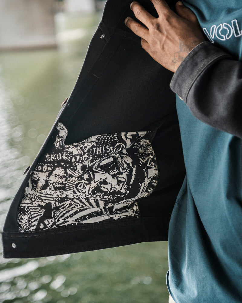 Wearing the Men’s Slakers Jacket by Volcom in black, a person opens the jacket to reveal a white graphic design featuring a stylized face and text printed on the inside. The background is blurred, with water visible behind them.