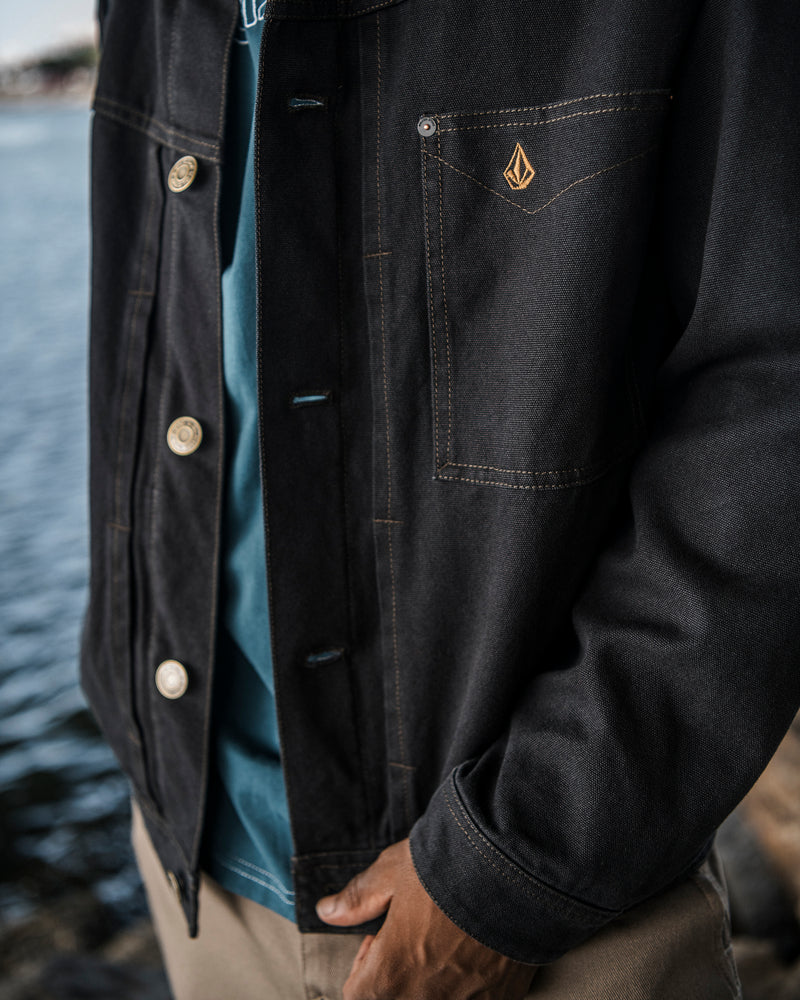A person stands by the water wearing a dark Volcom Men’s Slakers Jacket, a teal shirt, and beige pants. Only the torso and part of one arm are visible.