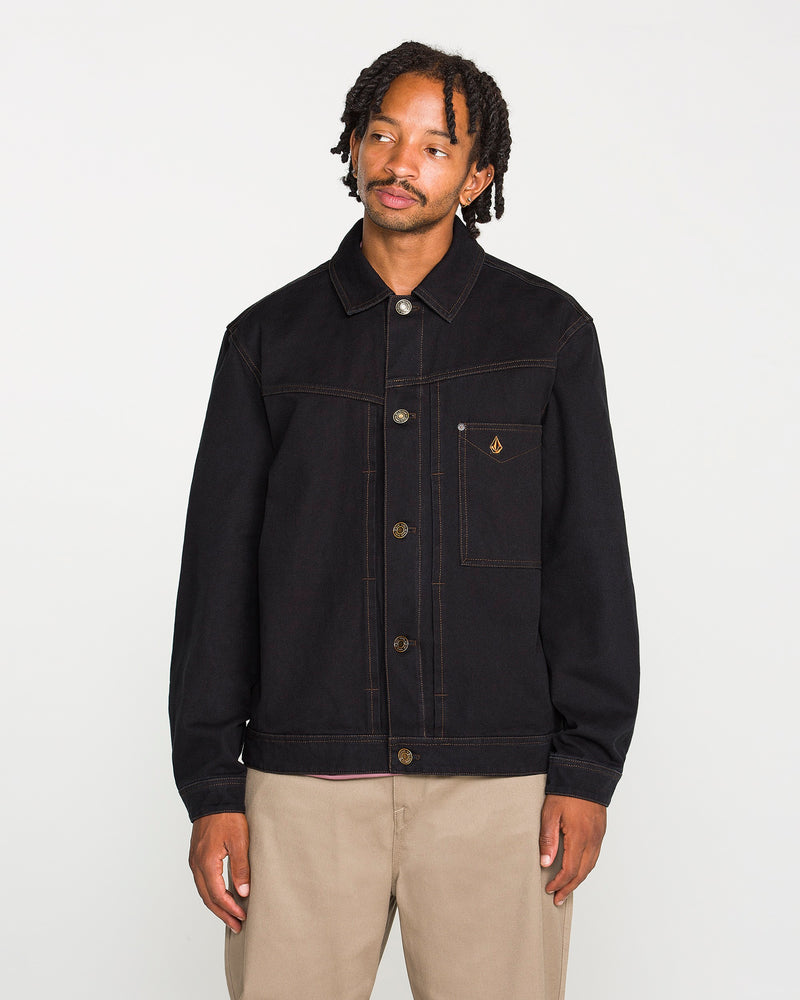 A person with short dreadlocks wears the Volcom Men's Slakers Jacket, a black lightweight piece featuring visible stitching and a small chest pocket, paired with beige pants while standing against a plain white background.