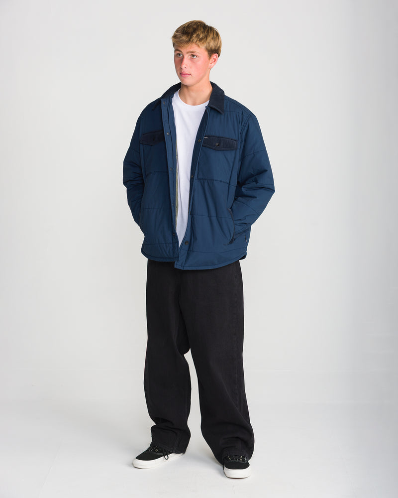 A person wearing a dark blue quilted Volcom Men’s Westshore Jacket with a corduroy collar over a white t-shirt and wide-leg black pants.