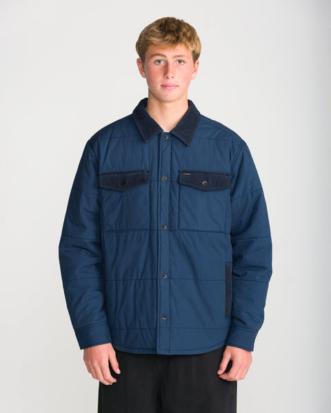 A person wears a Volcom Men’s Westshore Jacket, a dark blue quilted shirt jacket with a corduroy collar, featuring a button-down front and two chest pockets.