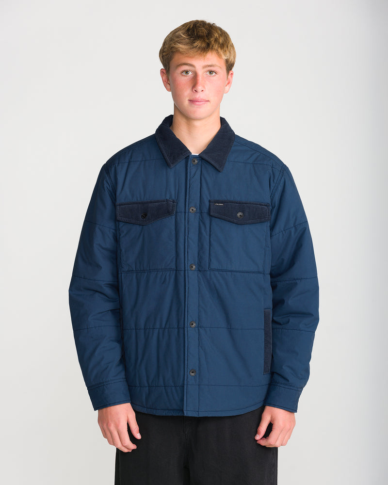 A person wears a Volcom Men’s Westshore Jacket, a dark blue quilted shirt jacket with a corduroy collar, featuring a button-down front and two chest pockets.