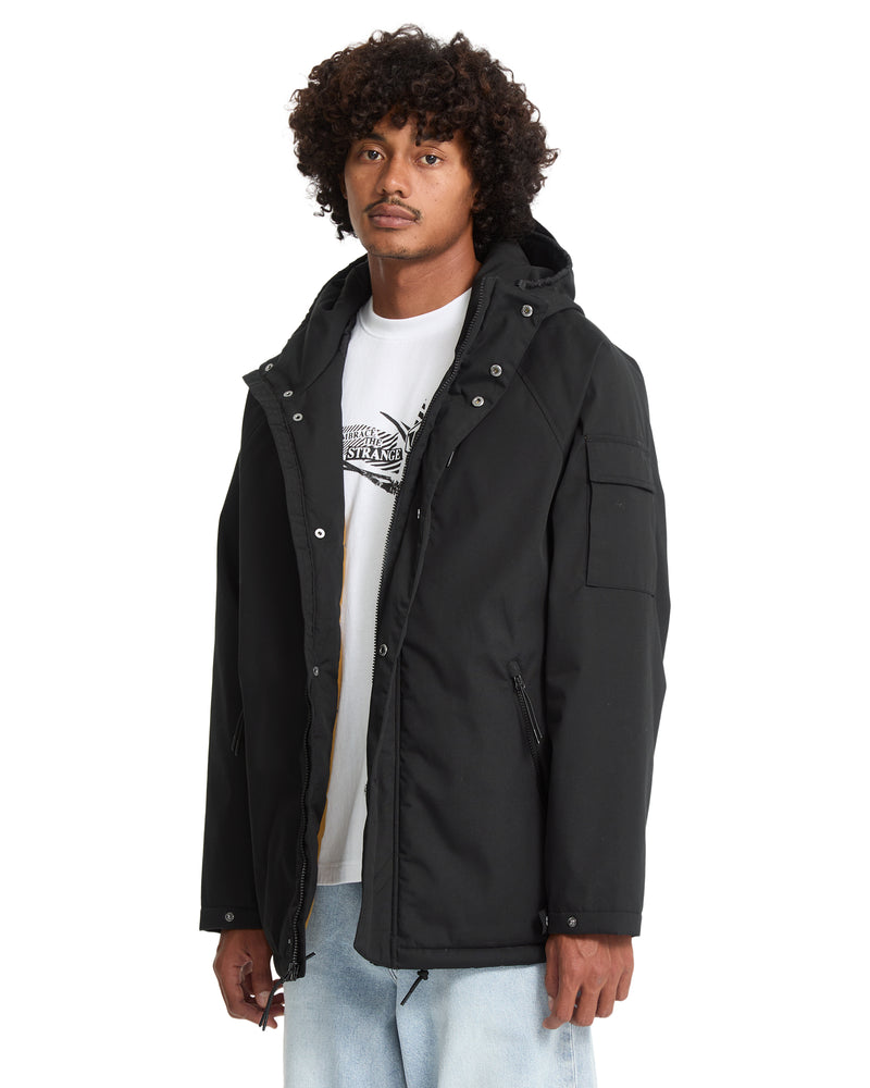 A person wears a black Volcom Men’s Stoke Stone II 10K Jacket, a water-resistant hooded jacket featuring a front zipper, snap buttons, and a utility pocket on the left sleeve.
