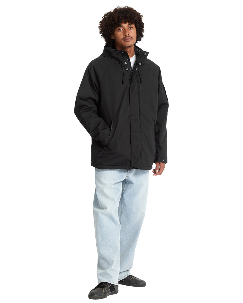 A man wears a Volcom Men’s Stoke Stone II 10K Jacket over a white t-shirt, paired with light-wash jeans and dark sneakers.