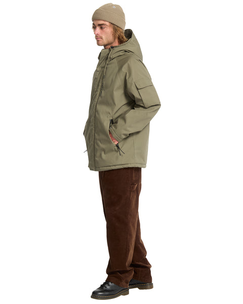 A man wears an olive green Volcom Men’s Stoke Stone II 10K Jacket with a hood and chest pocket, paired with brown corduroy pants and a beanie.