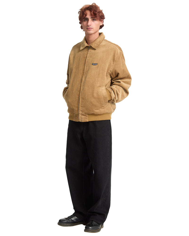 A person wearing a tan cotton corduroy Men’s Kurtis Jacket by Volcom with a blouson fit and zipper closure, paired with black trousers and black shoes.