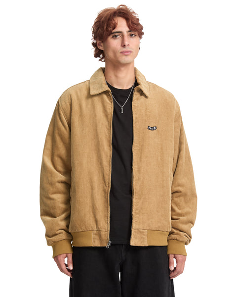 A man wears a tan cotton corduroy Volcom Men’s Kurtis Jacket in a blouson fit, featuring a collar and a small logo patch on the chest.