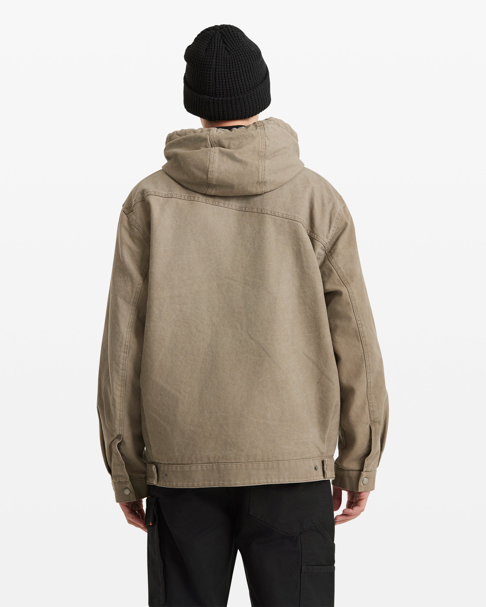 A person wears the Volcom Men's Freshdust Workwear Jacket in tan with a zip front, along with a black beanie and black pants, standing facing away from the camera against a plain white background.