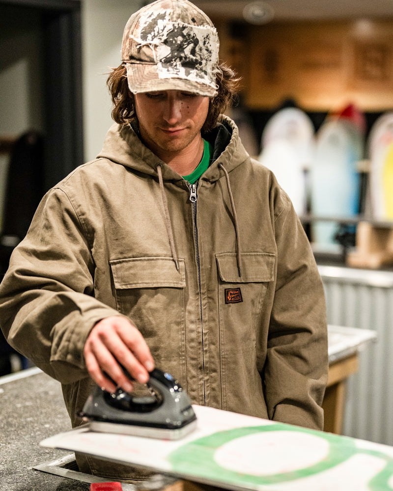 Wearing a tan Volcom Men’s Freshdust Workwear Jacket with a zip front closure and spacious hand and chest pockets, along with a camouflage hat, a person uses an iron on a snowboard in a workshop where surfboards are visible in the background.