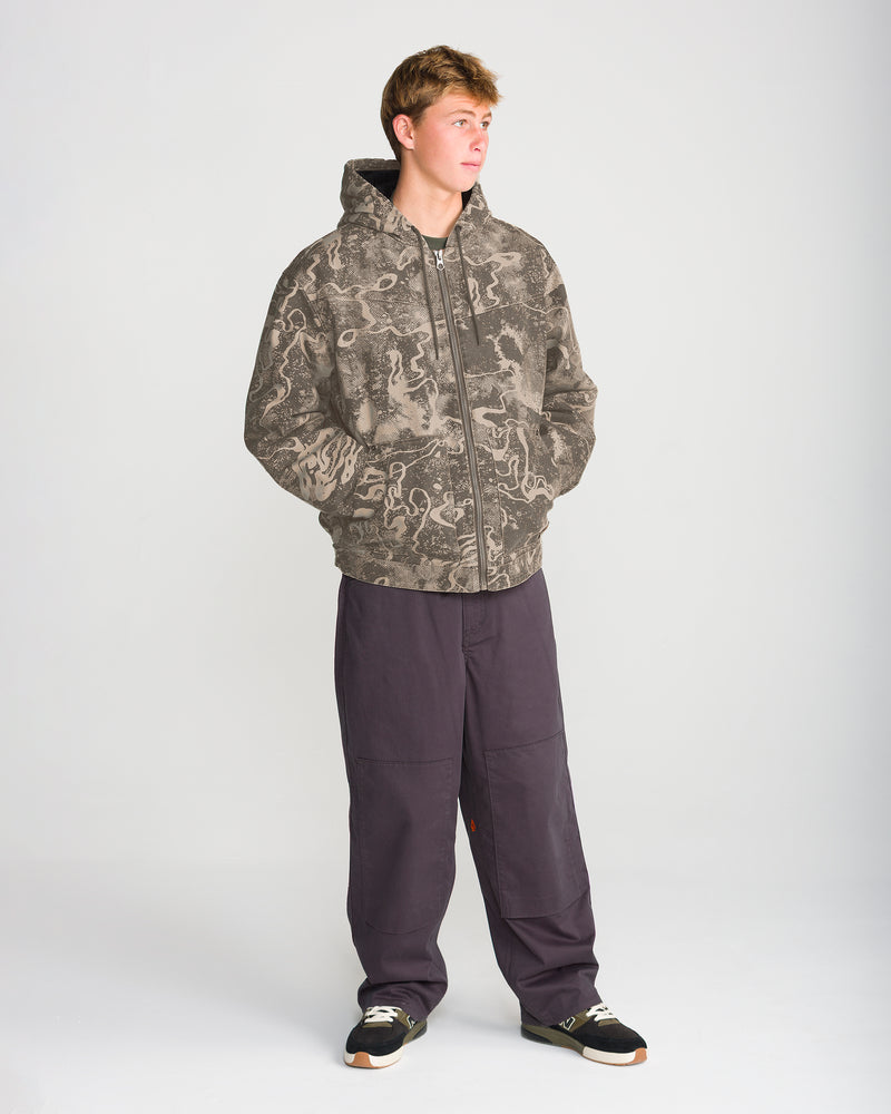 A person wearing a tan and brown marbled camo Volcom Men’s Fresh Storm Workwear hooded zip-up jacket, baggy dark grey pants, and black sneakers.