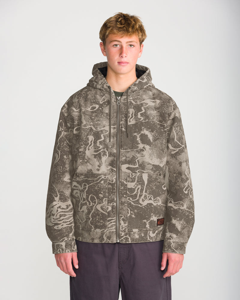 A person wears a Volcom Men’s Fresh Storm Workwear Jacket in a tan and brown abstract camo print over a dark shirt and pants.