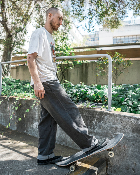 A man with a skateboard steps onto it by a concrete ledge outdoors, wearing a graphic tee, Volcom Men's Chillow Denim in baggy fit, and black shoes. He looks relaxed in the sunlight, surrounded by greenery.