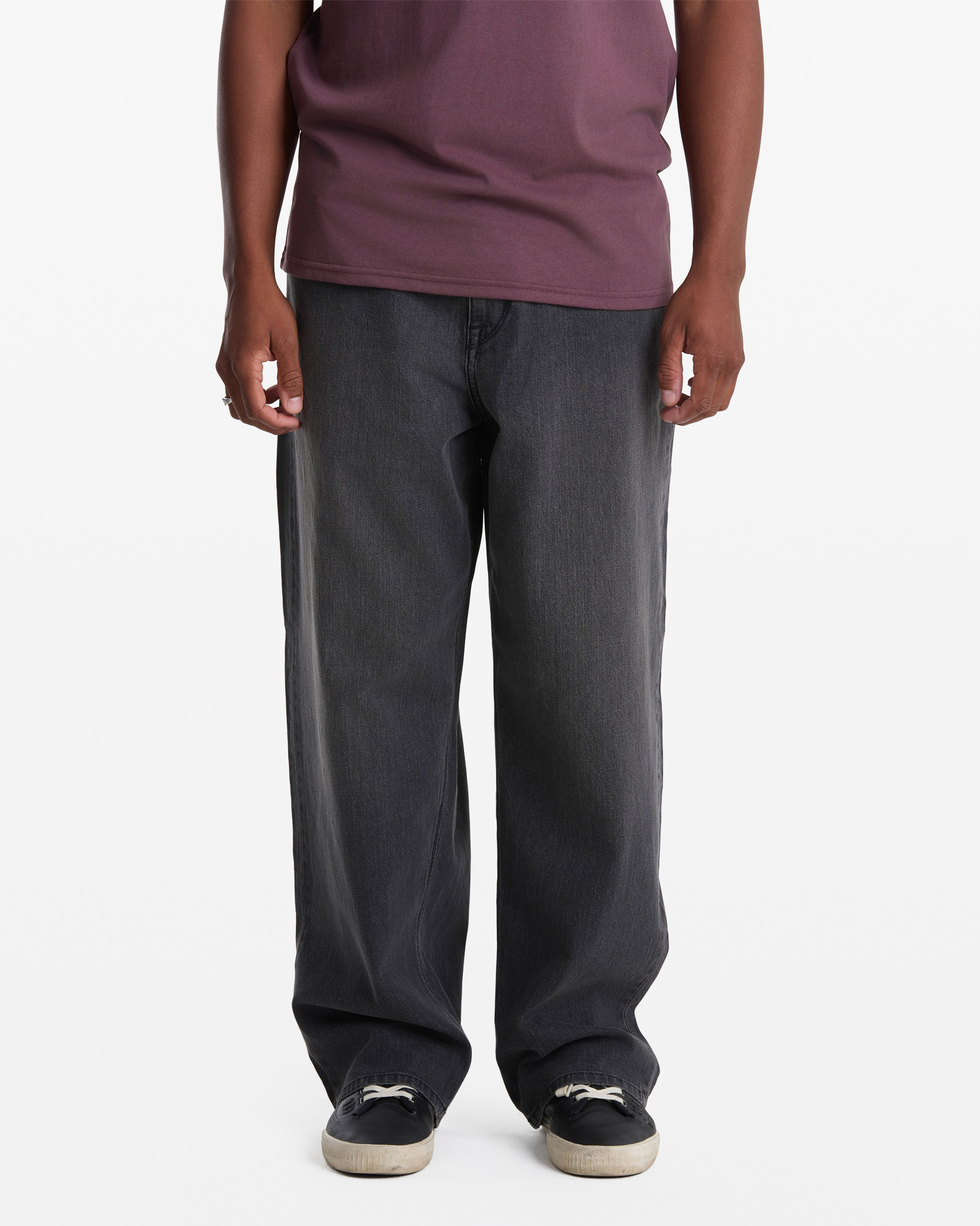 A person models Volcom's Men's Chillow Denim loose-fitting dark gray jeans with a maroon T-shirt and black sneakers against a white background. Their face is not visible.