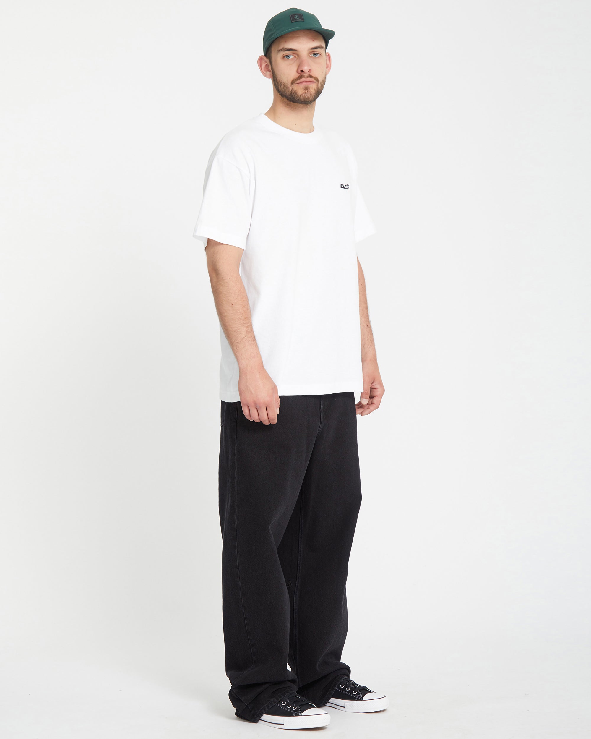 A man in a green cap, white t-shirt, Men's Chillow Denim jeans by Volcom Brand Jeans, and black sneakers stands against a plain white background, gazing to the side with a neutral expression.