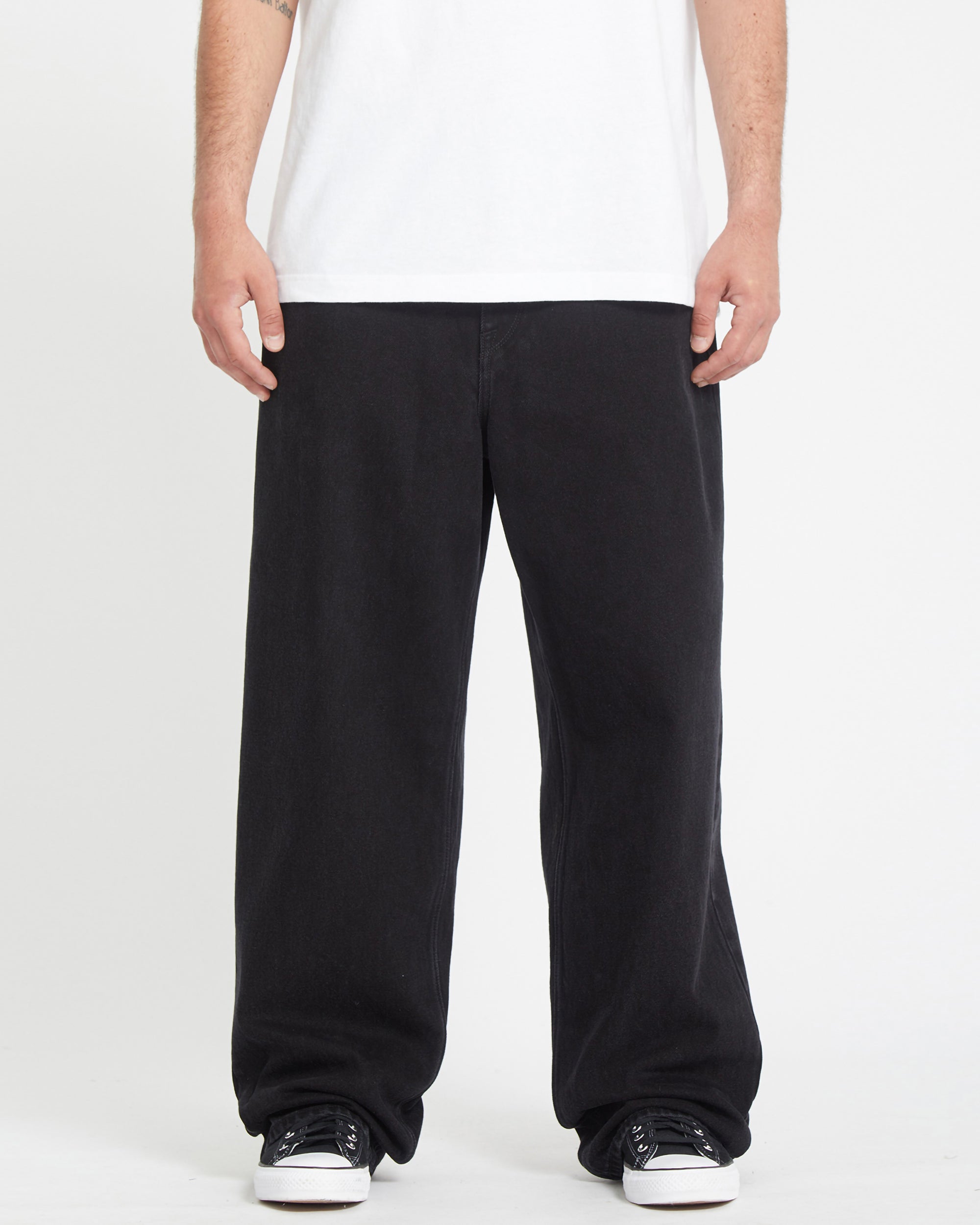 A person wearing a white T-shirt, Men's Chillow Denim baggy black jeans, and black-and-white sneakers stands against a plain white background with their face not visible.