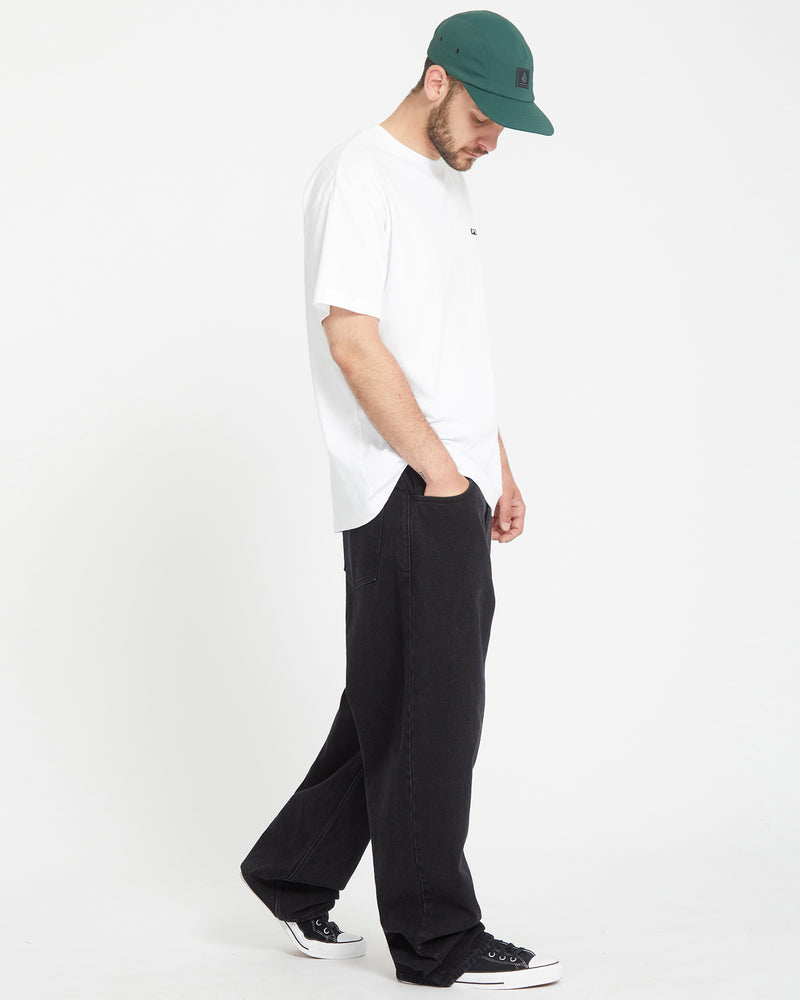 A man in a green baseball cap, white t-shirt, and baggy Men's Chillow Denim stands against a plain white background, looking down with his hands in his pockets.