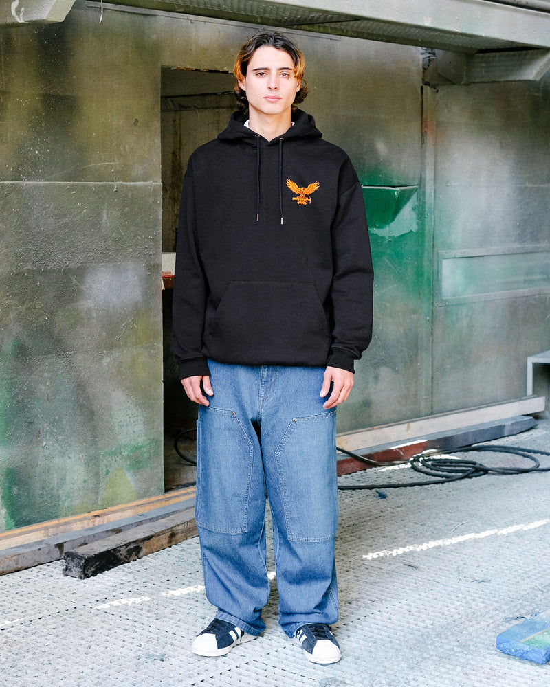 A person with shoulder-length hair, dressed in a black hoodie with a gold eagle emblem and Volcom Men's Work Carpenter Denim jeans, stands indoors on a textured floor with greenish walls and industrial details.