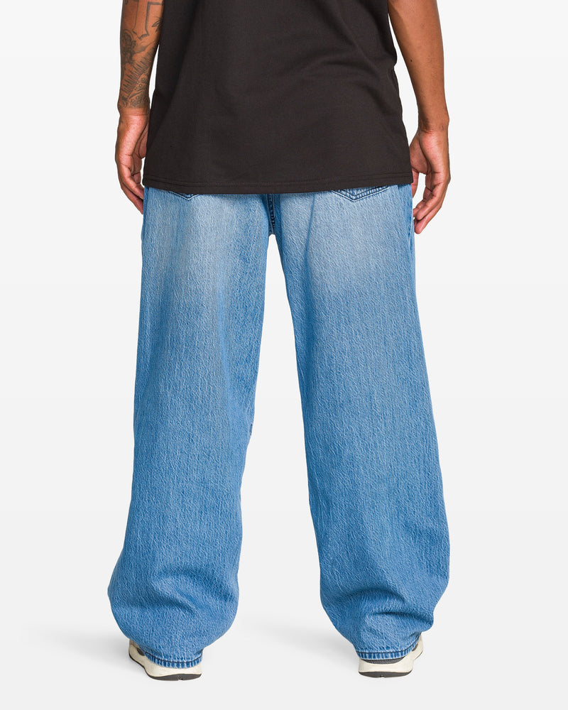 A person stands facing away, wearing a black T-shirt, Volcom Men's Chillow Denim Vs jeans with a baggy fit, and white sneakers. Their arms rest at their sides, revealing part of a tattoo on one forearm.