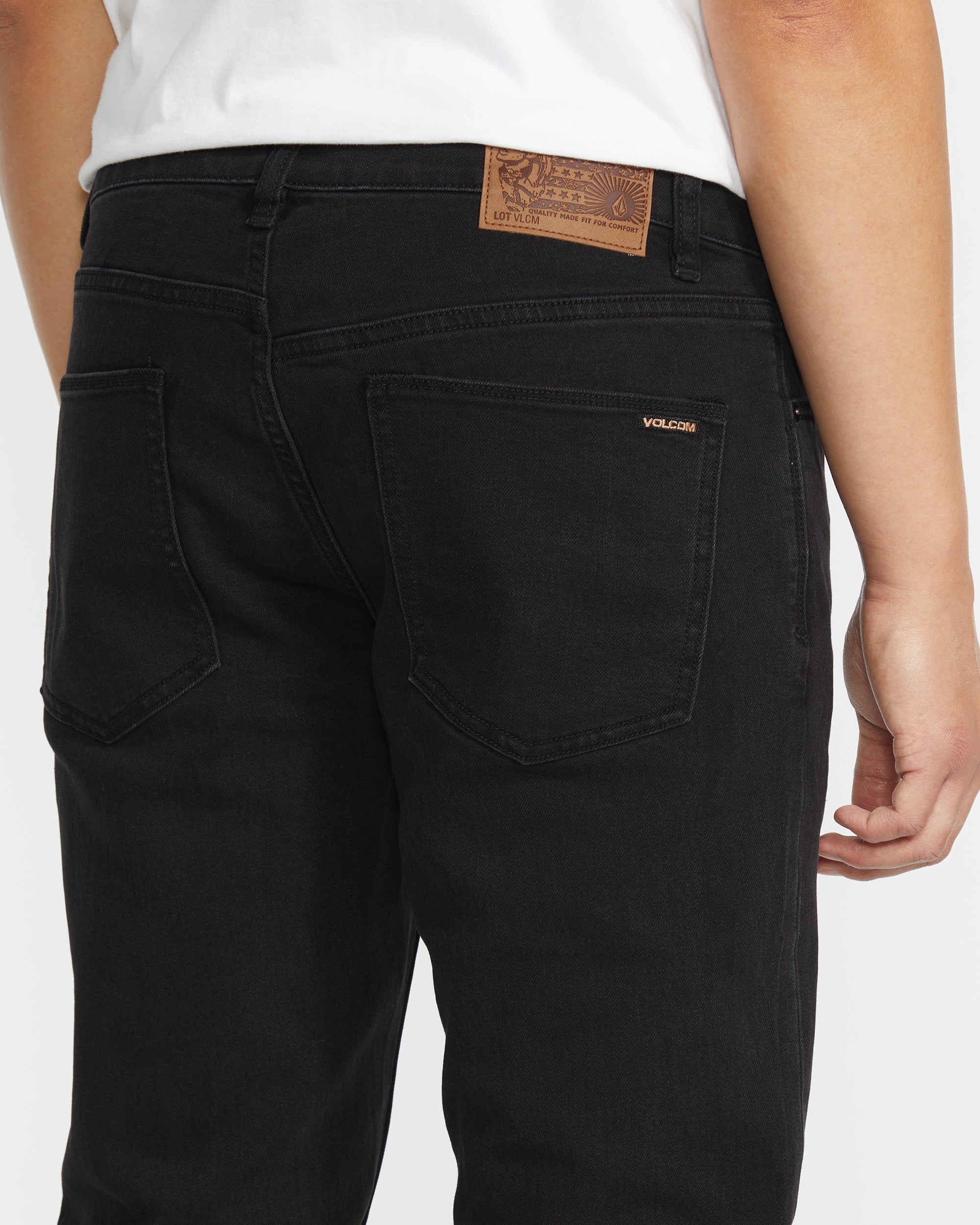 A person shown from the waist down wears black Vorta Denim slim fit jeans by Volcom made from stretch denim, paired with a white shirt. A brown leather patch and the Volcom brand name are visible above the right back pocket.