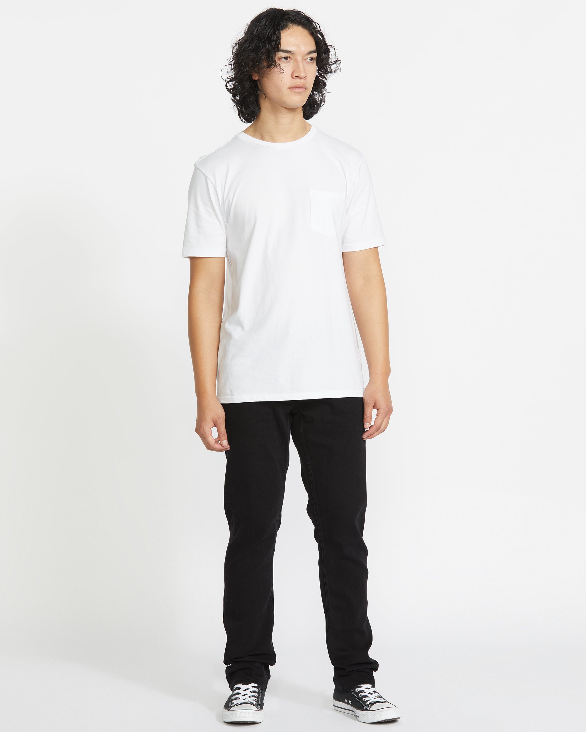 A person with curly dark hair stands against a white background, wearing a plain white t-shirt, black Volcom Vorta Denim straight leg jeans, and black low-top sneakers. They look slightly to the side with a neutral expression.