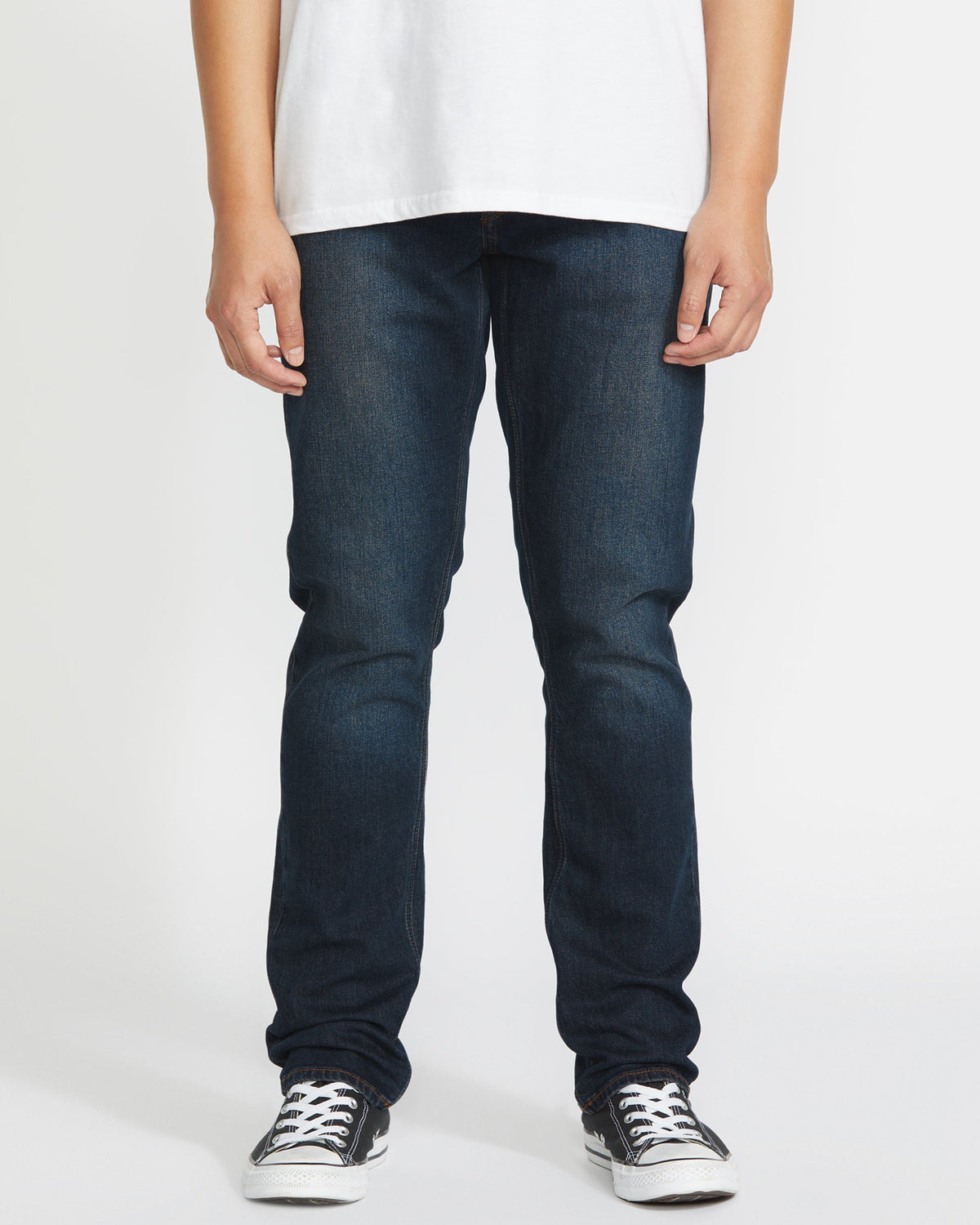 Wearing Volcom's Men's Vorta Denim jeans, a person stands against a plain white background. The image is cropped from the shoulders down and also shows a white t-shirt and black-and-white sneakers.