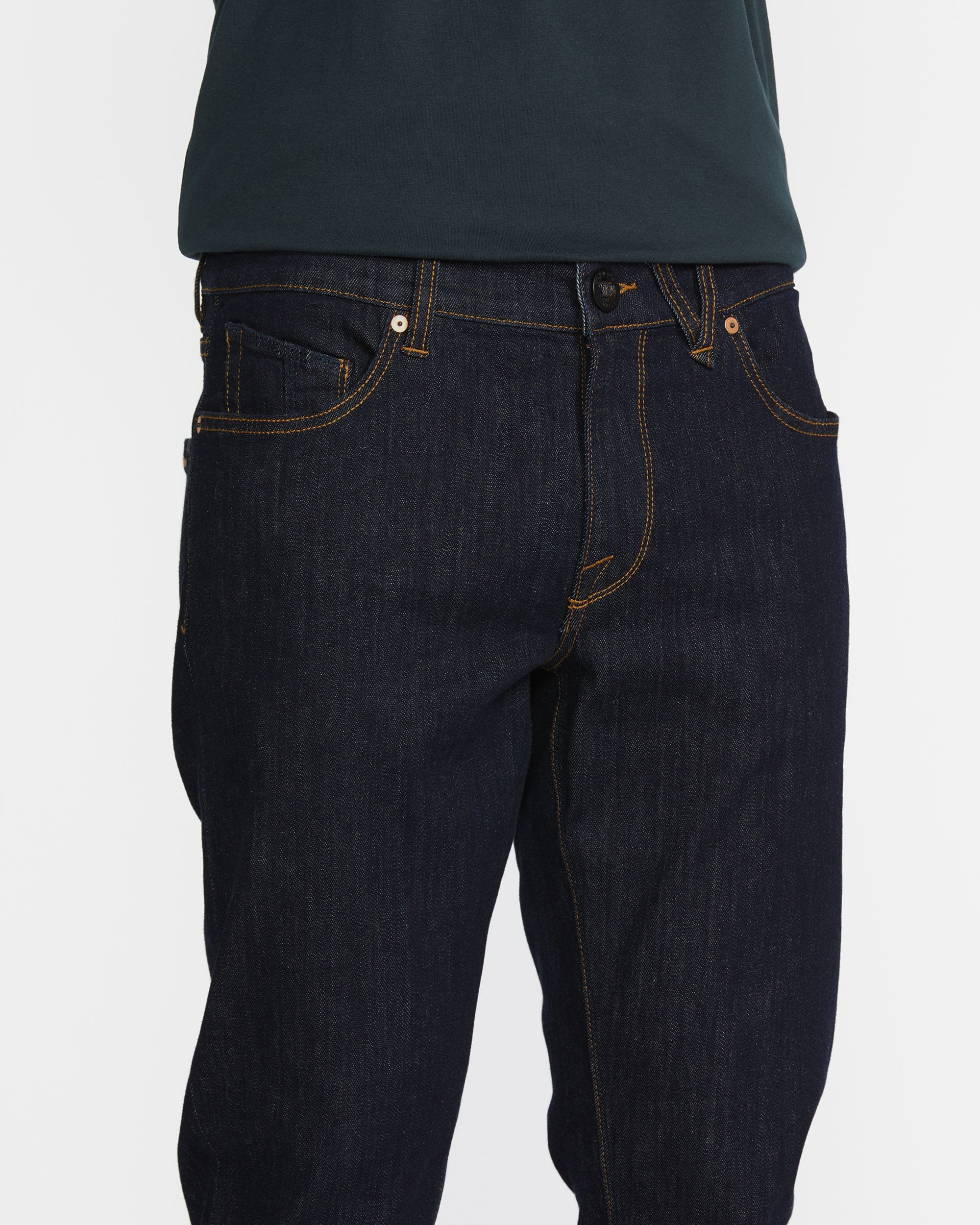 A person wears slim fit Men's Vorta Denim by Volcom with orange stitching and a dark green shirt. The photo focuses on the waist and upper legs against a white background, highlighting Water Aware details.