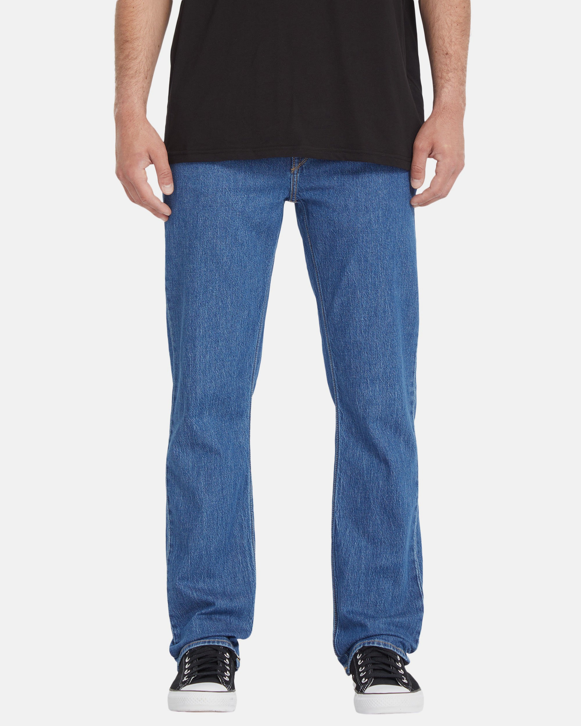 A person wearing a black t-shirt, Volcom Men's Solver Denim Pants in blue, and black-and-white sneakers, shown from the shoulders down against a plain white background.