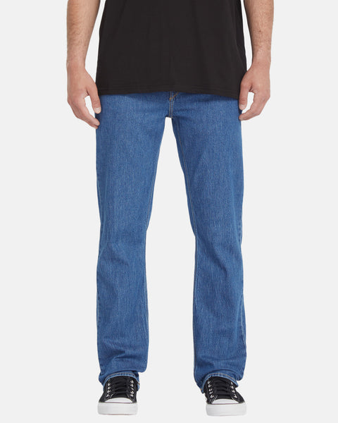 A person wearing a black t-shirt, Volcom Men's Solver Denim Pants in blue, and black-and-white sneakers, shown from the shoulders down against a plain white background.