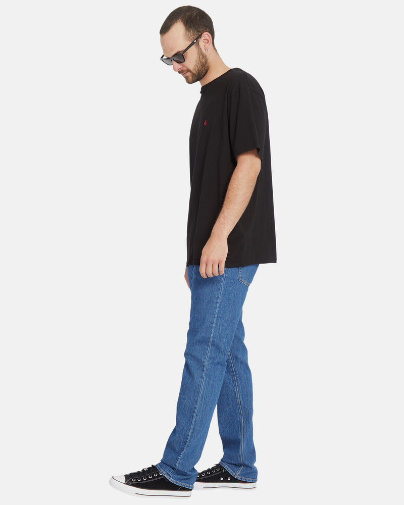 A man in sunglasses, a black t-shirt, Volcom Men's Solver Denim Pants, and black sneakers stands in profile against a plain white background.