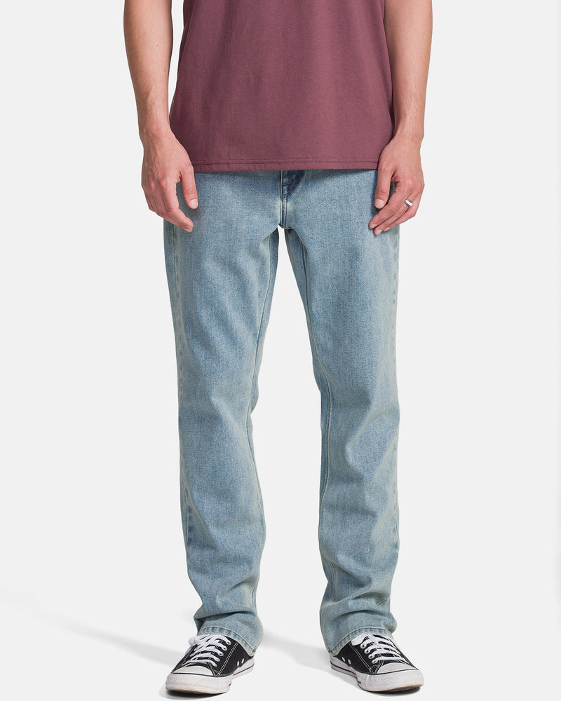 A person wearing a maroon t-shirt, Volcom Men's Solver Denim Pants in Dust Bowl Indigo, and black low-top sneakers stands against a plain white background. The image captures them from shoulders to feet.
