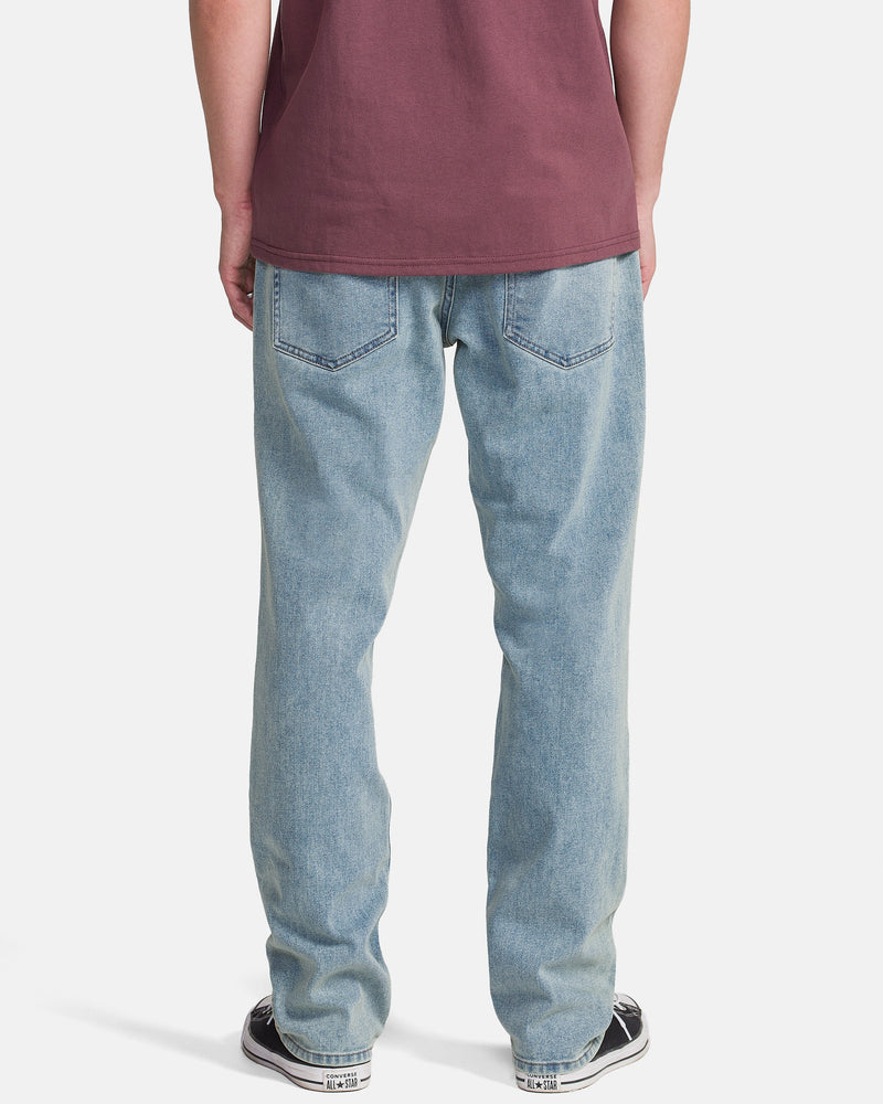 A person stands with their back to the camera, wearing Volcom Men's Solver Denim Pants in Dust Bowl Indigo, a maroon t-shirt, and black-and-white sneakers against a white background.