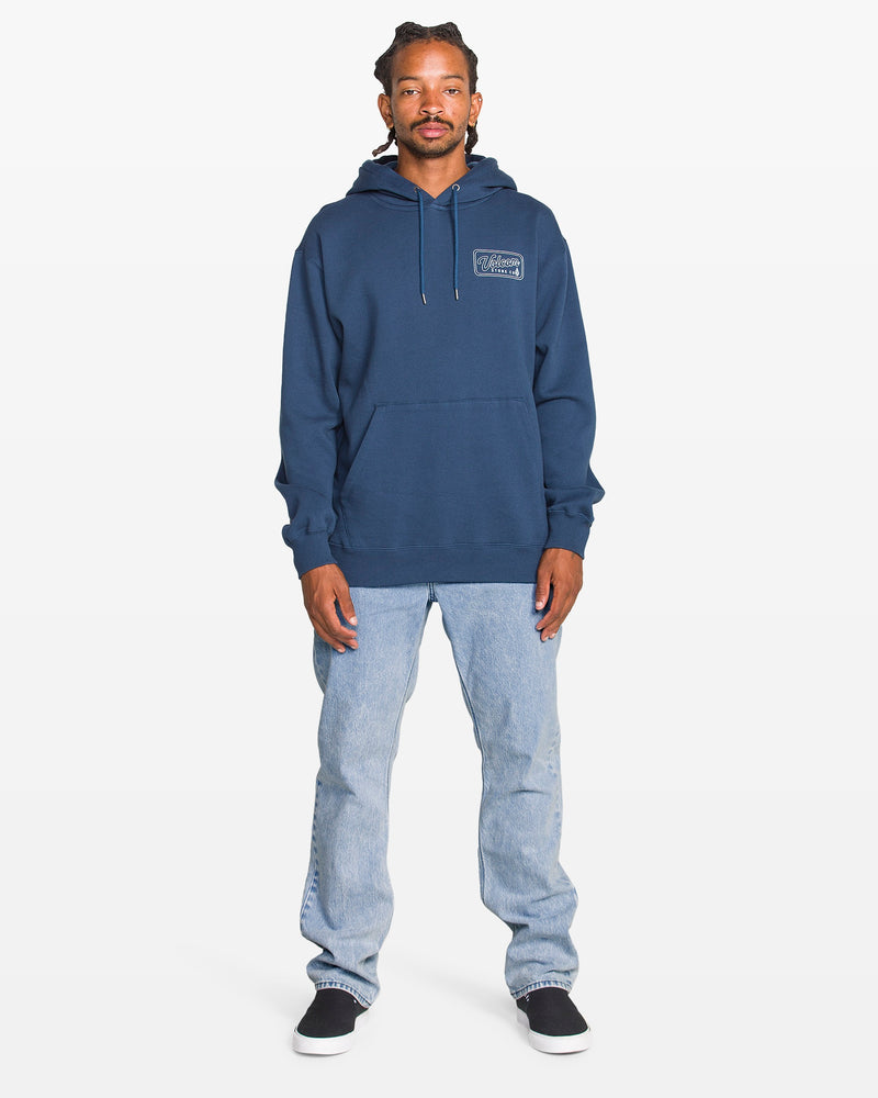 A man stands facing forward against a plain white background, wearing a blue hoodie, Volcom Men's Solver Denim Pants in a bleached-out indigo wash, and black slip-on shoes.