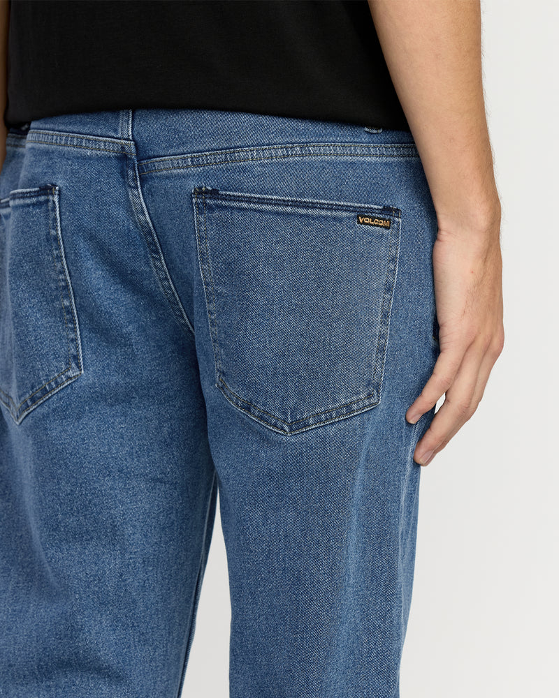 A person is shown from the waist down wearing Men's Solver Denim jeans in Deep River Blues by Volcom and a black shirt, with their right arm relaxed. The VOLCOM label is visible above the back pocket.